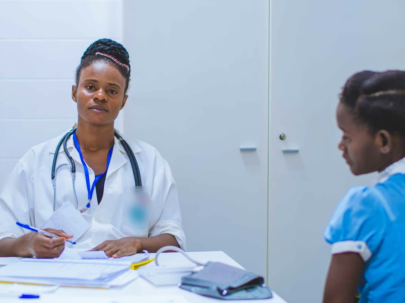 A clinician writing up notes at a consulting-room desk, with a child patient seated opposite. Paediatric and neonatal intensive care, for children and newborn babies, are planned services at TEMCO, opening 2029.